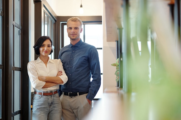 Portrait of happy smiling young businessman and businesswoman standing in office corridor and looking at camera