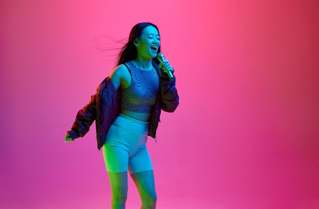 Portrait of happy smiling young girl singing in microphone against pink studio background in neon