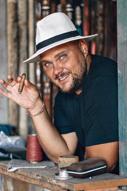 Portrait of a happy smiling young man with a beard and a black\
and white panama hat on his head, holding a cigar in his hand,\
leaning his logs on the table. summer concept.