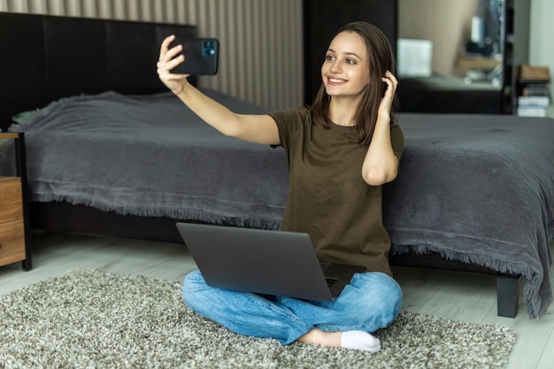 Ritratto di giovane donna sorridente felice sdraiata sul pavimento, usando il laptop e facendo selfie a casa