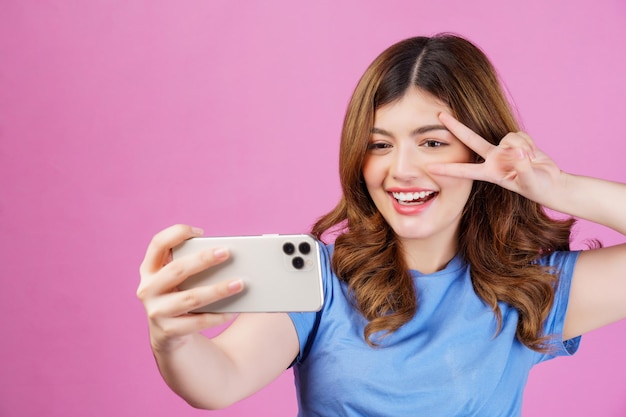 Портрет счастливой улыбающейся молодой женщины в повседневном селфи в футболке со смартфоном на розовом фоне