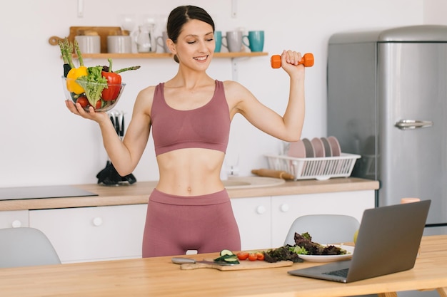 Portrait of happy sporty woman blogger holding a plate of fresh vegetables and showing biceps with dumbbells and leading video conference on healthy eating on laptop in kitchen