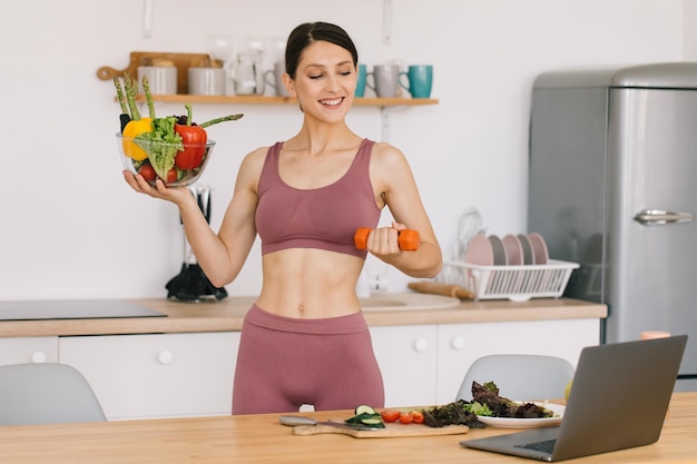 Portrait of happy sporty woman blogger holding a plate of fresh vegetables and showing biceps with dumbbells and leading video conference on healthy eating on laptop in kitchen