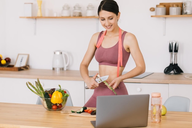 행복한 스포티 여성 블로거가 아스파라거스 요리를 하고 부엌에서 노트북에 대한 건강한 식생활 주제에 대한 화상 회의를 주도하는 초상화