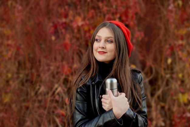 Portrait of happy student girl in red cap holding silver metal thermos in hand during autumn outside