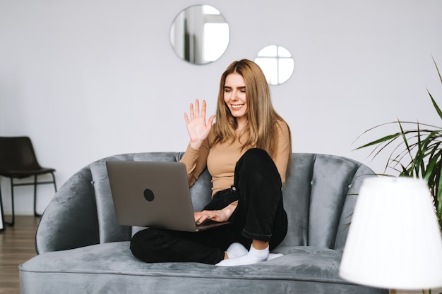 Ritratto di felice ed elegante giovane donna seduta su un divano, guardando lo schermo del laptop in videochiamata e saluto con agitando.