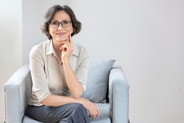 Portrait of happy, successful elderly woman with glasses sitting in a chair, smiling aged mature woman in stylish clothes looking at camera