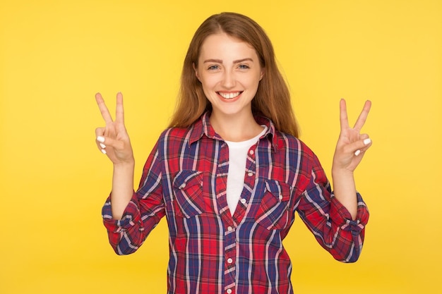 勝利のジェスチャーをし、勝利に自信を持って笑顔で幸せな成功した生姜の女の子の肖像画は、黄色の背景で隔離の屋内スタジオショット指で平和のサインを示しています