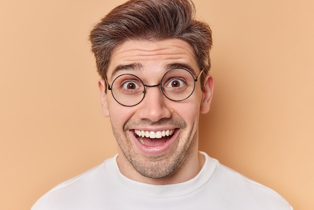 Photo portrait of happy surprised adult man reacts on amazing news wears round transparent eyeglasses smiles broadly dressed in casual wear isolated over brown background human reactions concept