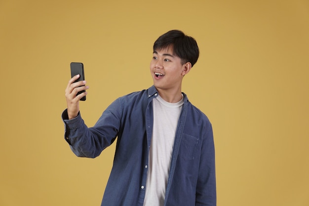 Portrait of happy surprised excited young asian man dressed casually looking at smartphone isolated