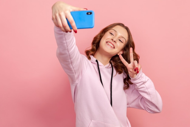 Portrait of happy teen girl in hoodie showing victory gesture with fingers on smartphone camera and sincerely smiling vlog or selfie Indoor studio shot isolated on pink background