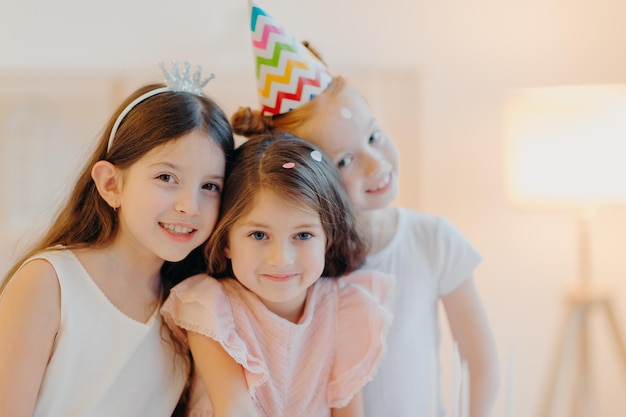 Portrait of happy three friends wear festive clothes party hat\
pose indoor against white background play together during birthday\
celebration cute girls have good mood come on special occasion