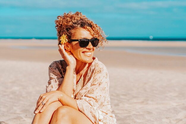 Portrait of happy tourist sitting and smiling at the beach with blue sky and ocean in background Travel and tourism in summer holiday vacation Female people with sunglasses on the sand