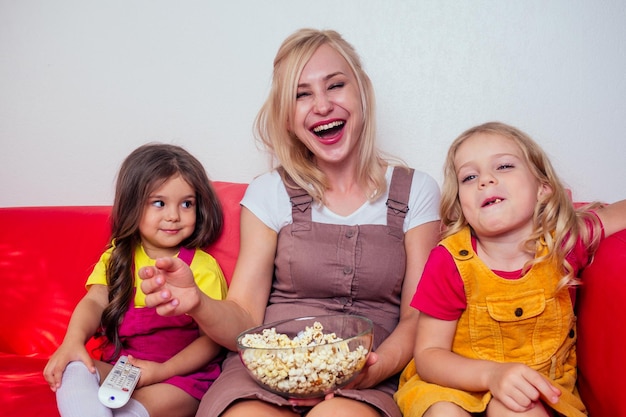 Portrait of happy two daughter and mother watching cartoons with popcorn on a red couch