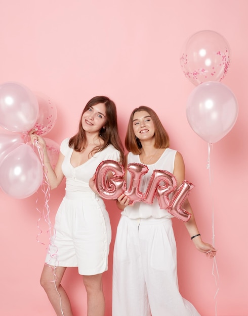 Photo portrait of happy two young women dressed in a white holdings a balloons isolated pink background