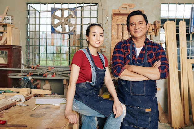 Portrait of happy Vietnamese carpenters in denim aprons standing in workshop