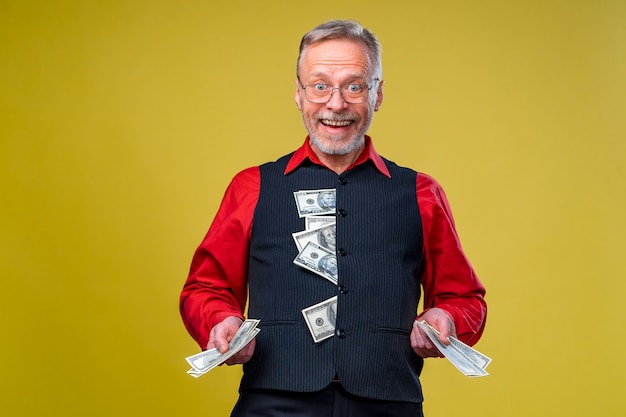 Портрет счастливой и белой улыбки зубов старший старый деловой человек, держащий деньги в руках, одетый в красную рубашку, изолированную на желтом фоне. Человеческие эмоции и выражения лица