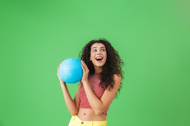 Portrait of happy woman 20s wearing summer clothes smiling and\
holding volley ball while standing on green