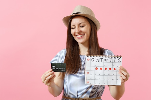 Ritratto di donna felice in abito blu, cappello con carta di credito, calendario dei periodi, controllo dei giorni delle mestruazioni isolati su sfondo rosa di tendenza. concetto ginecologico sanitario medico. copia spazio.