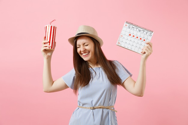 Portrait of happy woman in blue dress, hat holding cup of soda, female periods calendar for checking menstruation days isolated on pink background. Medical healthcare gynecological concept. Copy space