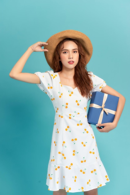 Portrait of a happy woman in dress and summer hat holding present box isolated over blue background