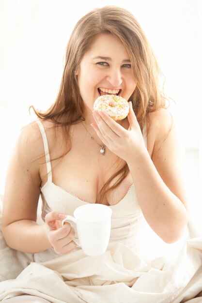 Portrait of happy woman eating donut and drinking coffee in bed