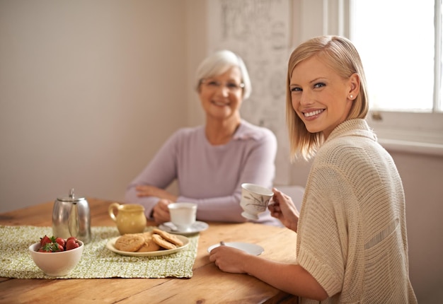 Portrait happy woman and elderly mother drinking tea at breakfast bonding and smile in house Face senior mom and daughter with coffee cup at table food and family eating cookies at home together