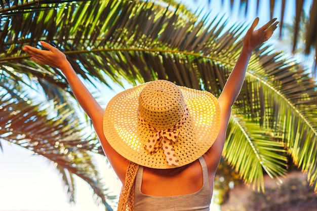 Portrait of happy woman in hat lifting hands under palm leaf in hotel yard Summer vacation Traveling