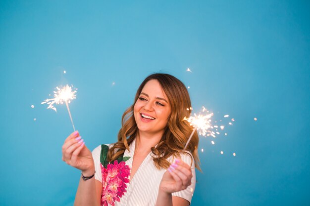 Portrait of a happy woman holding bengal lights over blue background. Christmas, celebrations and holidays concept.