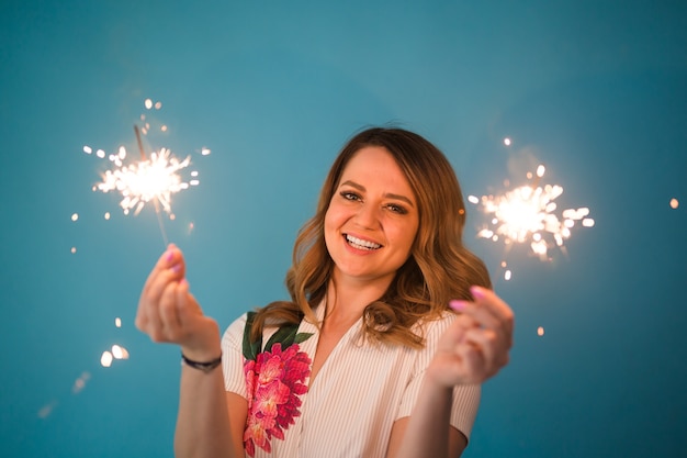 Portrait of a happy woman holding bengal lights over blue background with copy space. Christmas, celebrations and holidays concept