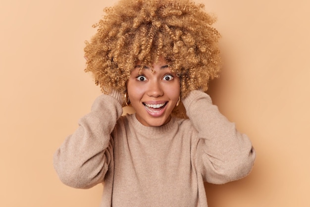 Photo portrait of happy woman looks with surprised expression grabs head giggles positively reacts on awesome news dressed in jumper isolated over beige background gets incredible excellent opportunity