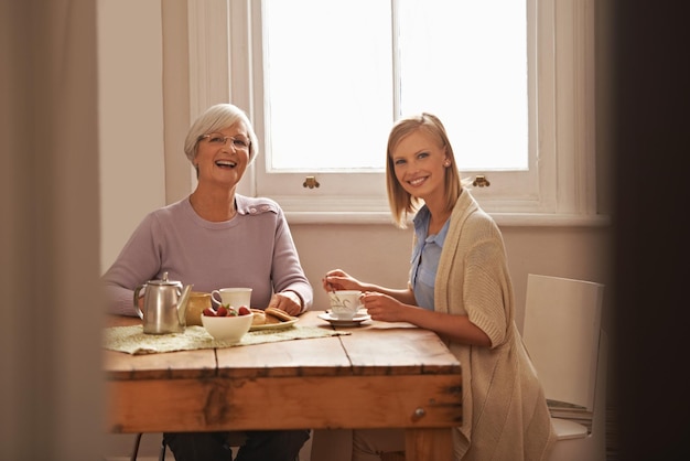 Portrait happy woman and senior mother drinking tea at breakfast bonding and smile in house Face elderly mom and daughter with coffee cup at table food and funny family laughing at home together