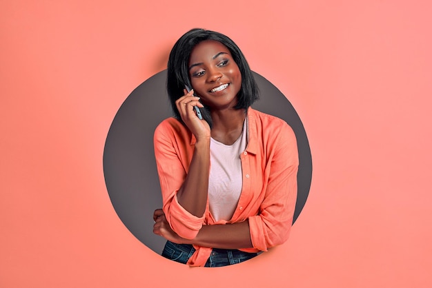 Portrait of happy woman talking on mobile phone in a circle hole in a coral background Copy space