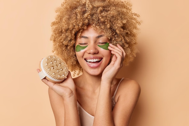 Portrait of happy woman touches face gently applies green hydrogel patches for skin refreshment holds body brush keeps eyes closed stands bare shoulders isolated over beige studio background