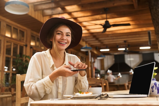 スタイリッシュなカフェに座っている間、携帯電話で食べ物を撮影する帽子をかぶって幸せな女性の肖像画