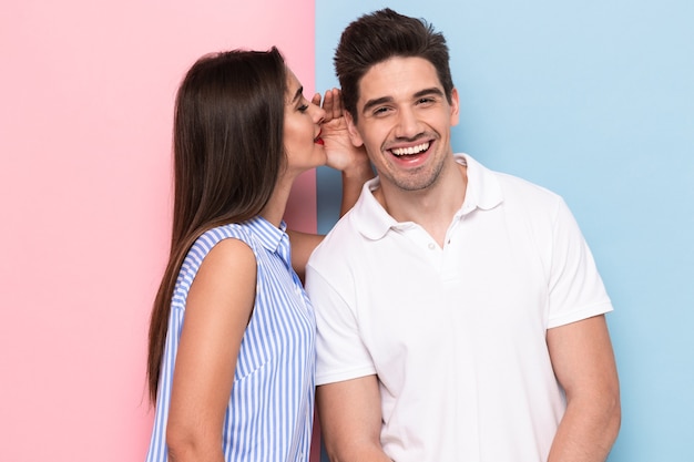 Portrait of happy woman whispering secret or interesting gossip to handsome man in his ear, isolated over colorful wall