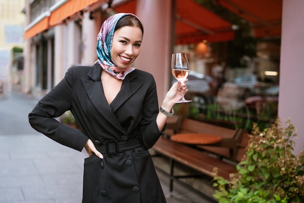 Portrait of a happy woman with makeup stands near a cafe on the street with a glass of wine in her hand