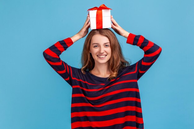 Портрет счастливой женщины с волнистыми волосами в полосатом повседневном свитере, стоящей с завернутой подарочной коробкой над головой и смотрящей в камеру. Крытая студия снята на синем фоне.