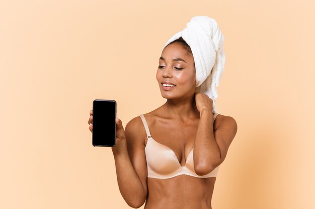 Portrait of happy woman wrapped in towel and wearing white lingerie using cell phone, isolated over beige wall