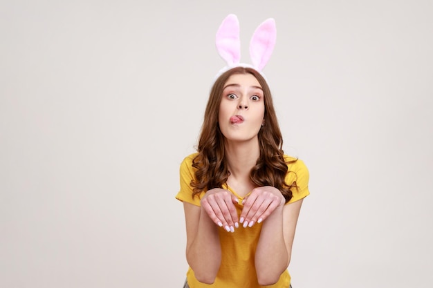 Portrait of happy woman of young age in yellow T-shirt with bunny ears and making hare paws with hands, childish grimace, humorous expression. Indoor studio shot isolated on gray background.