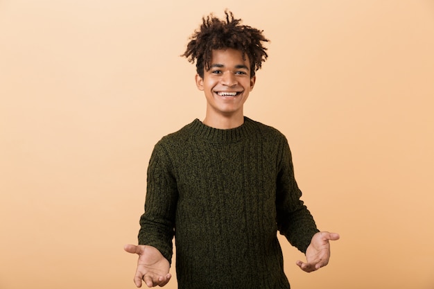 Portrait of a happy young africa man dressed in sweater isolated over beige wall, outstretched hands