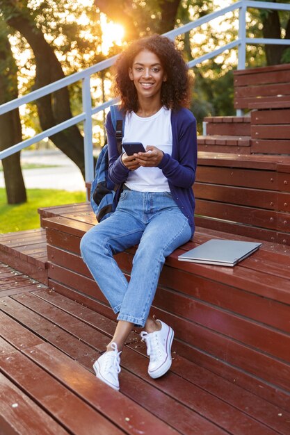 Ritratto di una giovane ragazza africana felice con lo zaino utilizzando il telefono cellulare mentre si riposa al parco