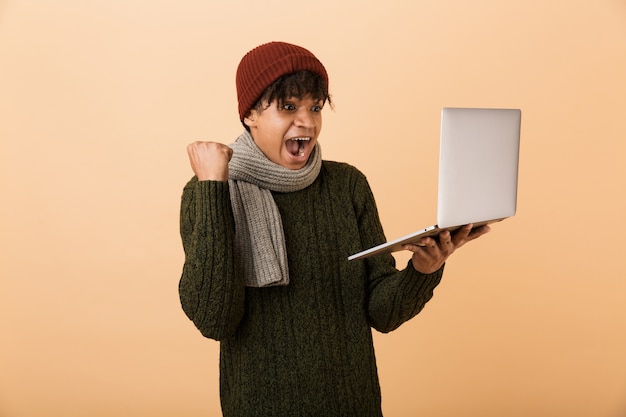 Ritratto di un felice giovane africano vestito in abiti autunnali isolati su muro beige, tenendo il computer portatile, celebrando