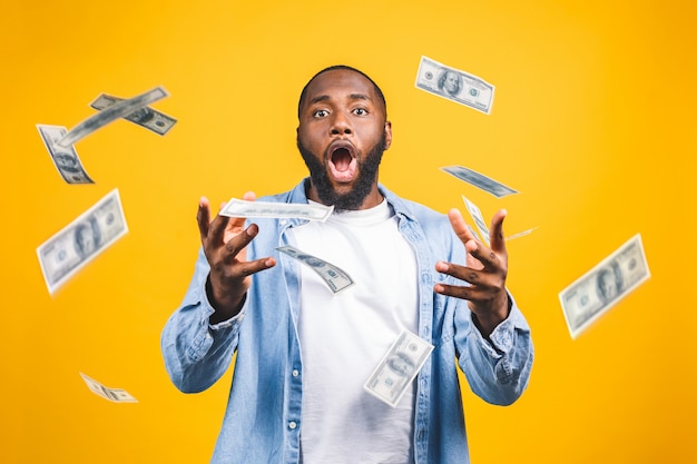 Portrait of a happy young afro american man throwing out money banknotes isolated over yellow background