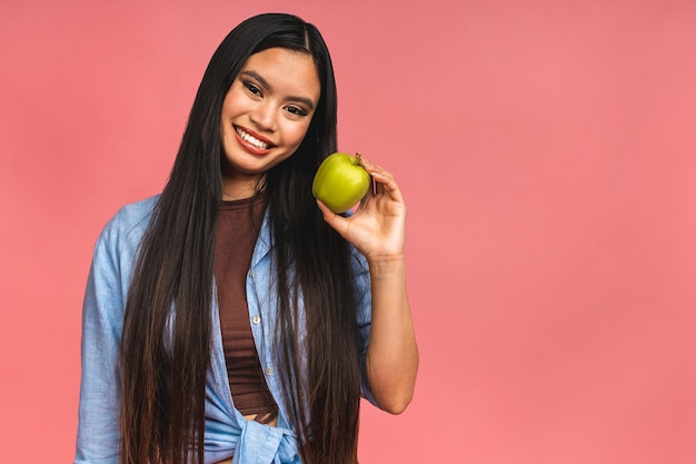 Portrait of a happy young asian chinese korean japanese woman holding or eating green apple isolated over pink background