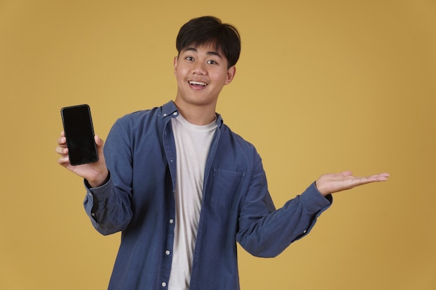 分離されたスマートフォンの空白の白い画面を表示してさりげなく笑顔を着て幸せな若いアジア人男性の肖像画