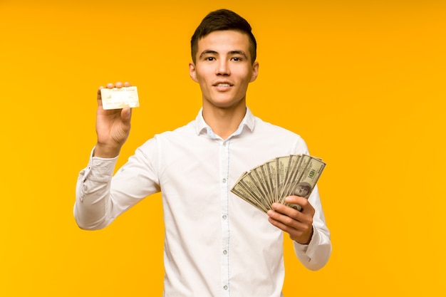 Portrait of happy young asian man holding credit card and money in hand smiling and looking at camera on isolated yellow background