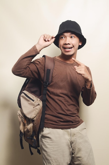 Portrait of happy young Asian man pointing at empty space wearing black hat carrying backpack for travel isolated on background