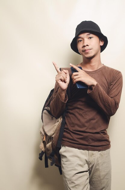 Portrait of happy young Asian man pointing at empty space wearing black hat carrying backpack