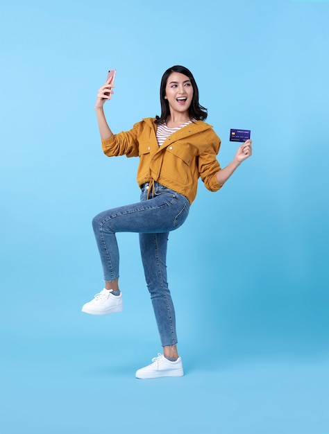 Portrait of a happy young asian woman celebrating with mobile phone and credit card over blue.